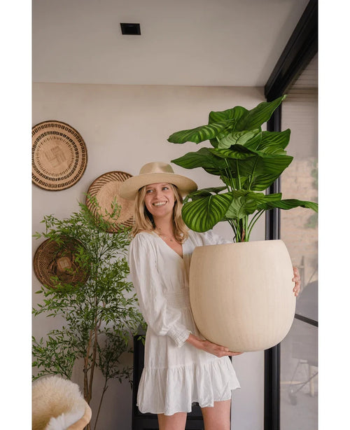 En kvinna i vit klänning och hatt ler medan hon håller den artificiella Calathea UV 72 cm-växten inomhus, med vävda korgar och en annan växt i bakgrunden.