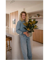 En kvinna med lockigt blont hår, klädd i jeanskläder, står och ler i ett modernt kök med växten Artificiell Magnolia i en 46 cm kruka i händerna, omgiven av träskåp och hyllor – ett idealiskt val för alla som letar efter växter med lite underhåll för att förbättra sin stilfulla interiör.
