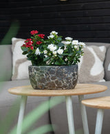 En Artificiell Plant Patio Rose 35 cm kräm UV står på ett runt träbord framför en beige soffa med mönstrade kuddar.