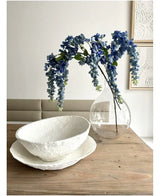 Ett blomsterarrangemang från Kunst Wisteria på 115 cm står i en genomskinlig vas på ett träbord, bredvid två vita skålar med struktur, medan väggen bakom är dekorerad med abstrakt konst.