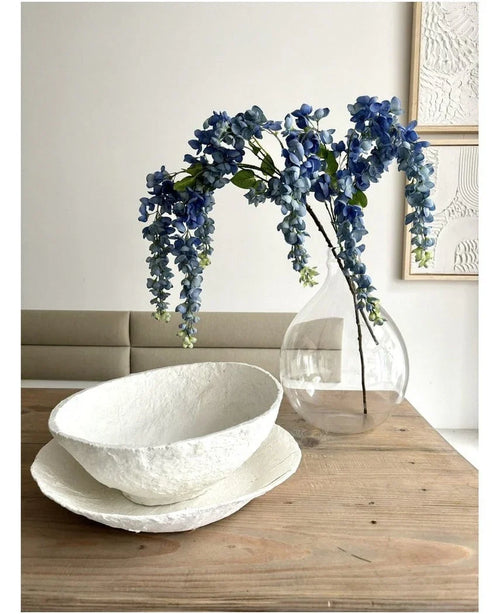 Ett blomsterarrangemang från Kunst Wisteria på 115 cm står i en genomskinlig vas på ett träbord, bredvid två vita skålar med struktur, medan väggen bakom är dekorerad med abstrakt konst.