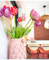 En vas med rosa hjärtmotiv håller Artificiell Blomma French Tulip 60 cm lila på ett matbord med stolar med vävda ryggstöd. I bakgrunden bidrar en rund spegel och en brunmönstrad vägg till den mysiga, moderna inredningen.