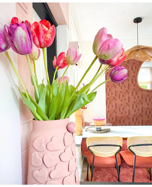 En rosa vas med präglade hjärtan håller den konstgjorda blomman fransk tulpan 60 cm röd på ett matbord. Bakom den står stolar med flätad rygg, en tårta på bordet och en spegel på en mönstrad brun vägg under en vävd hänglampa.