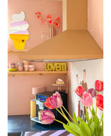 Ett pastellrosa kök med Artificiell Blomma French Tulip 60 cm lila i förgrunden, en gul LOVETTI tallrik på hyllan, rosa muggar, väggkonst av en glassstrut och en minimalistisk väggmålning av två personer på väggen.