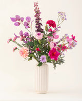 En vit texturerad vas innehÄller Purple Dawn siden konstgjorda blombuketten i rosa, lila och lavendel nyanser med grönt bladverk, mot en ljus, neutral bakgrund.