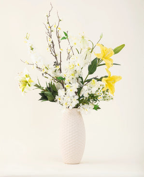 En vas med vit struktur visar elegansen hos Sunkissed Ivory siden konstgjorda blombuketten, med gula liljor, vita blommor och gröna blad mot en vanlig ljus bakgrund.