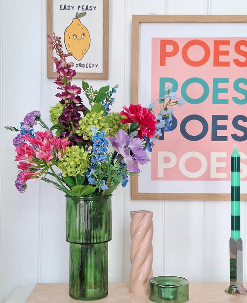 En grön glasvas rymmer en konstgjord sidenblommabukett Blossom Breeze pÄ bordet. Bakom den hÀnger inramade bilder - en med citron och "EASY PEASY", en annan med "POESY" - vilket ger extra charm. I nÀrheten finns ett grönt ljus och ett rosa spiralljus frÄn Blossom Breeze.