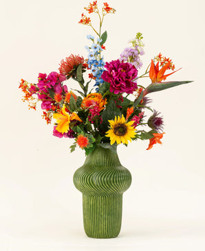En grön strukturerad vas visar den konstgjorda blombuketten Colorful Spirit i siden, med solrosor, rosa pioner, orange och lila blommor och frodig grönska mot en vanlig ljus bakgrund.