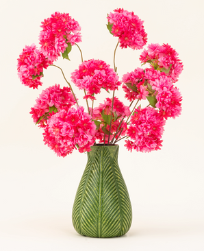 En grön vas med bladmotiv visar den konstgjorda sidenblommabuketten Cherry Blossom Pink - ljusrosa, fluffiga silkeskörsbärsblommor med gröna blad - mot en vanlig ljus bakgrund.
