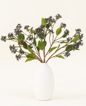 En bukett med konstgjorda sidenblomster Japanese Berry Steel Blue med långa stjälkar, mörklila bär och stålblad står i en vas med vit struktur mot en vanlig ljus bakgrund.