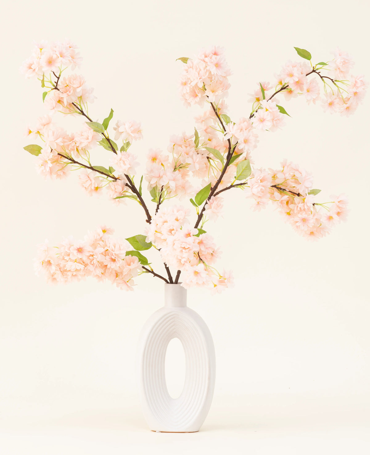 En vit keramikvas med en bred oval öppning visar flera grönbladiga sidenkonstgjorda blombuketter Körsbärsblommor ljusrosa grenar mot en enkel ljus bakgrund.