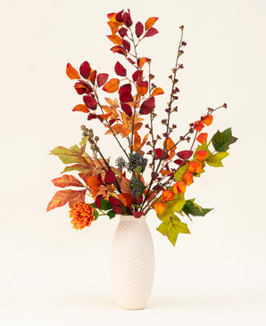 En vas med vit textur visar upp Chestnut Charm Silk Artificiell blombukett, med höstlöv, orange blommor och grönt blad - ett elegant konstgjort arrangemang mot en vanlig ljus bakgrund.