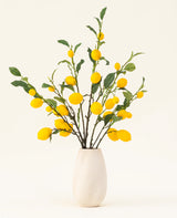 En vas med vit textur visar Lemons konstgjorda blombukett i siden, med gröna blad och livfulla gula citroner mot en ljus bakgrund.