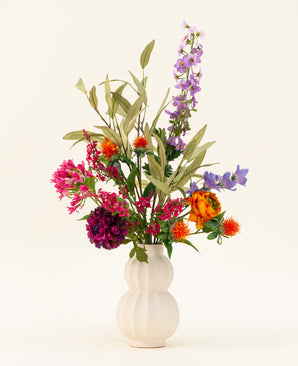 En vit, texturerad vas visar upp den färgglada Svenski konstgjorda sidenblommabuketten, med livfulla lila, orange, rosa och blå konstgjorda blommor med gröna blad som lyser upp alla utrymmen mot en enkel ljus bakgrund.