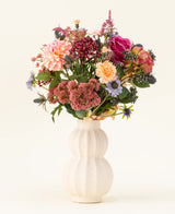 I en vit, böjd keramikvas finns den konstgjorda blombuketten Pretty Pastel siden - en färgstark blandning av hållbara blommor som rosa rosor, dahlior, lila och orange blommor och grönska - vackert arrangerad mot en ljus bakgrund.