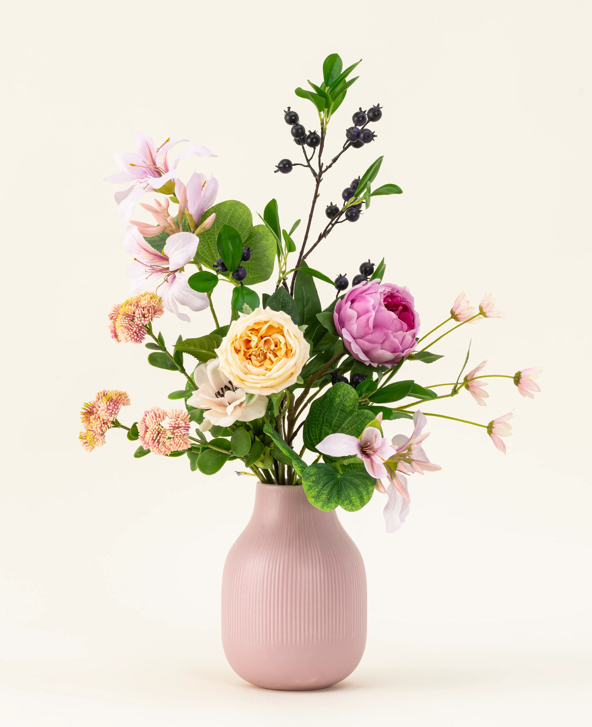 The Touch of Cloudlight konstgjord blombukett i siden, presenterad i en rosa keramikvas, innehåller rosa pioner, gula rosor, små blommor, grönska och grenar med mörka bär mot en ljus bakgrund.