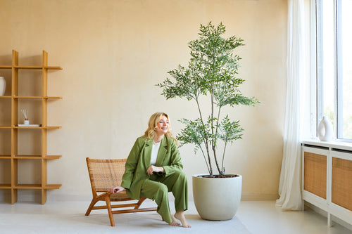 En kvinna i grön kostym ler när hon sitter på en trästol bredvid växten Artificiell Ruscus 180 cm i ett ljust, modernt rum med beiga väggar, stora fönster och minimalistisk inredning.