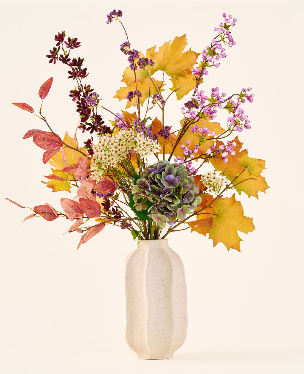 'Wild Amethyst'-buketten med konstgjorda sidenblommor, med höstlöv, lila och vita blommor och gröna hortensior, är i en vit keramikvas mot en enkel, ljus bakgrund.