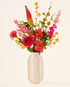 I en vit keramikvas finns 'Summer Blush'-buketten med konstgjorda sidenblommor, bestående av rosa, gula och orangea blommor med gröna blad, mot en vanlig ljus bakgrund.