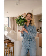 En leende kvinna i jeansoutfit håller den Artificiella Gardenia UV 39 cm vit konstgjord växt i ett modernt kök med träpallar, vita bänkskivor och neutral inredning.