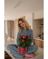 En kvinna med lockigt blont hår, iklädd jeansoverall och rosa sneakers, sitter med benen över kors på en köksbänk och ler medan hon håller i den Artificiella Gardenia UV 39 cm-växten i fuchsia.