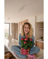 En leende kvinna med lockigt blont hår sitter i kors på en ljus, modern bänkskiva och håller i Artificiell Gardenia UV 39 cm fuchsiaväxt – en rosa krukväxt med livfulla röda blommor. Hon är klädd i jeans, blå skjorta och rosa skor.