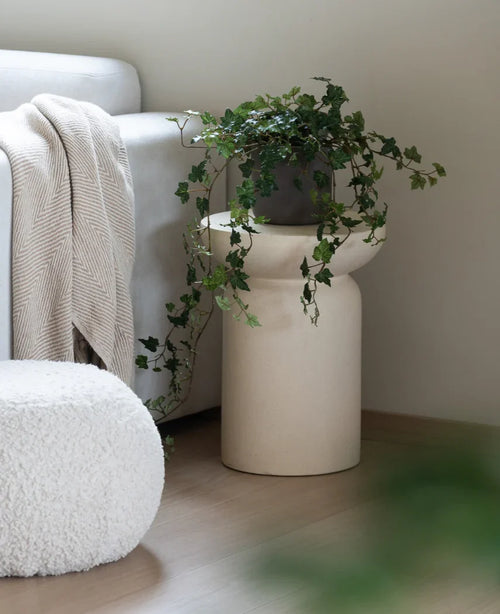 En Artificial Ivy Deluxe Green 90 cm står i en beige vas på ett trägolv bredvid en ljusgrå soffa med en krämfärgad pläd och en vit strukturerad puff.