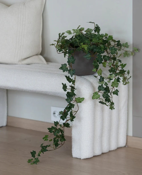 Artificial Ivy Deluxe Green 90 cm, med sina frodiga hängande rankor, står på en vit strukturerad bänk bredvid en krämfärgad kudde i ett minimalistiskt rum med ljusa trägolv.