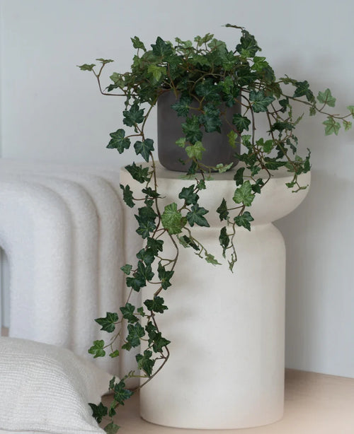En Artificial Ivy Deluxe Green, en 90 cm hängande murgröna i en grå kruka, står på en modern, vit, skulpturell bas bredvid en vikt vit filt och en vit strukturerad kudde.