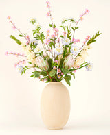 I en vas med beige struktur ligger Silk konstgjorda blombuketten Lots of Love XL, en handbunden bukett av rosa, vita och blå sidenblommor med gröna blad och långa stjälkar mot en ljus bakgrund.