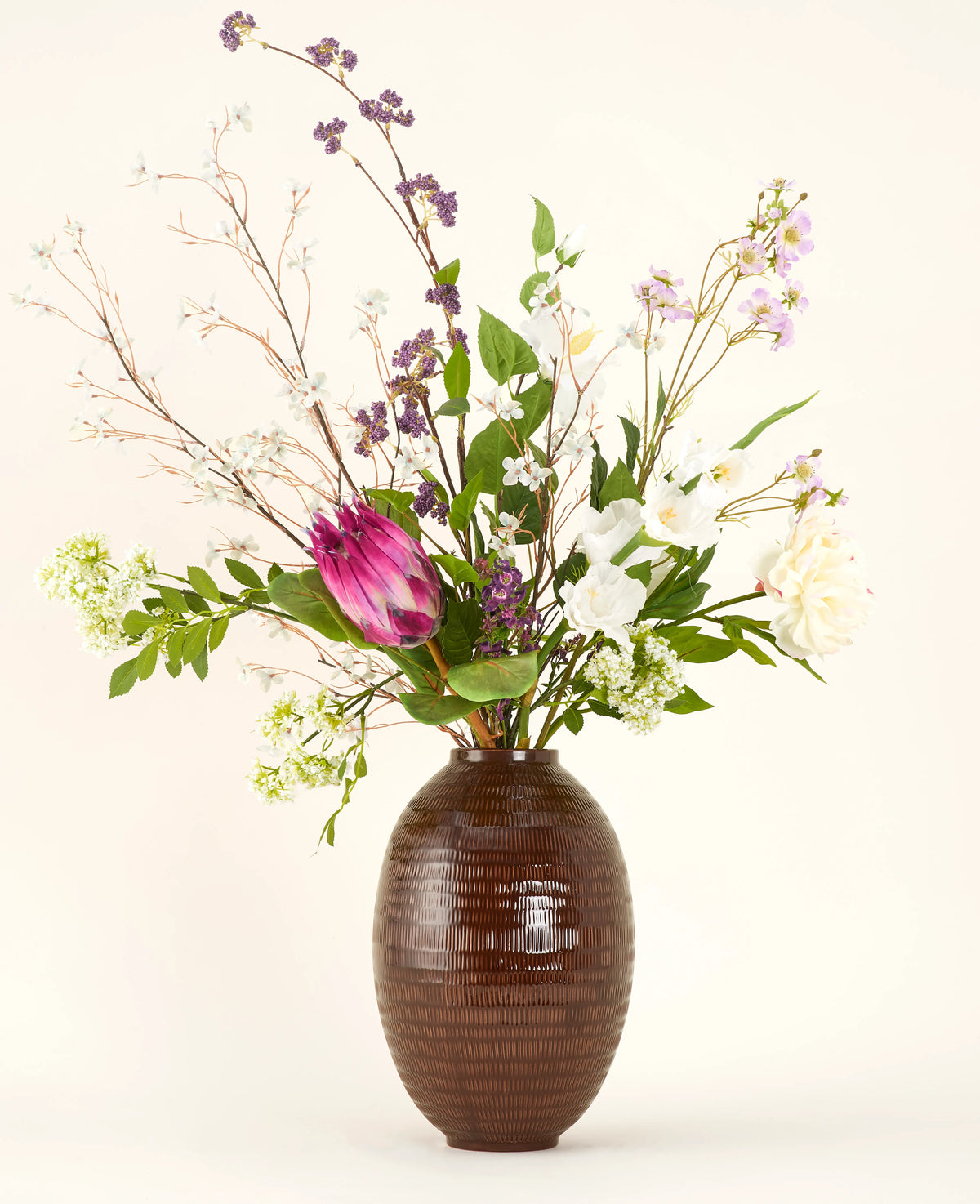 En Serene Bloomfield konstgjord blombukett i siden, med en stor rosa protea, vita rosor, lila och vita blommor och grönt bladverk, står i en brun räfflad vas mot en vanlig ljus bakgrund.
