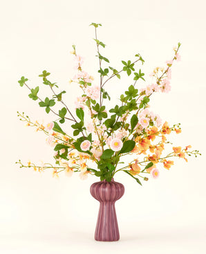 En konstgjord blombukett i Blossom Harmony siden är arrangerad i en lila vas med rosa och persikablommor och gröna blad, mot en enkel ljus bakgrund för en elegant visning.