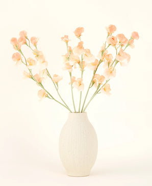 Den krämfärgade keramikvasen innehåller Silk konstgjorda blombuketten Pea Peach, med delikat persika och ljusrosa blommor med långa gröna stjälkar mot en ljus, neutral bakgrund.