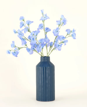 En blå räfflad keramikvas visar den konstgjorda blombuketten Pea Blue siden med sina delikata ljusblå konstgjorda blommor mot en enkel, ljus bakgrund.
