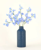 En blå räfflad keramikvas visar den konstgjorda blombuketten Pea Blue siden med sina delikata ljusblå konstgjorda blommor mot en enkel, ljus bakgrund.