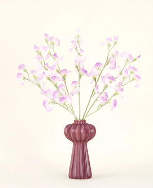 Silk konstgjorda blombuketten Pea lilac har långa stjälkar av ömtåliga, ljuslila blommor och gröna blad i en lila keramisk vas med vertikala åsar, mot en vanlig, benvit bakgrund.