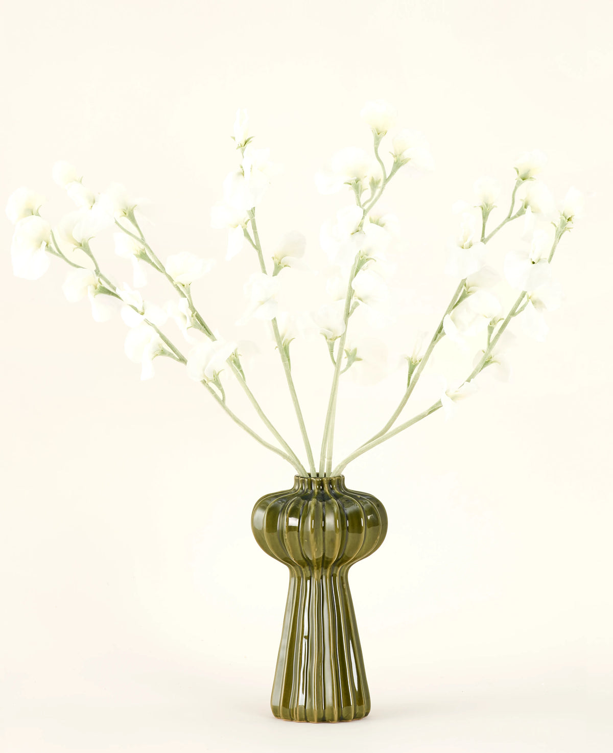 En grön räfflad glasvas håller Silk konstgjorda blombuketten Pea cream med fina vita sidenblommor och smala stjälkar, mot en vanlig benvit bakgrund.