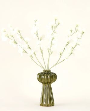 En grön räfflad glasvas håller Silk konstgjorda blombuketten Pea cream med fina vita sidenblommor och smala stjälkar, mot en vanlig benvit bakgrund.