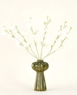 En grön räfflad glasvas håller Silk konstgjorda blombuketten Pea cream med fina vita sidenblommor och smala stjälkar, mot en vanlig benvit bakgrund.