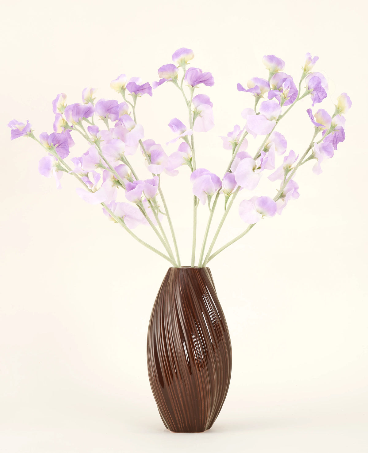 En texturerad brun vas rymmer den konstgjorda sidenblommabuketten Pea lila-ljuslila sidenblommor med grönaktiga stjälkar mot en enkel, ljus bakgrund.
