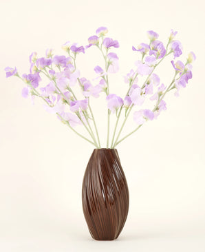 En texturerad brun vas rymmer den konstgjorda sidenblommabuketten Pea lila-ljuslila sidenblommor med grönaktiga stjälkar mot en enkel, ljus bakgrund.