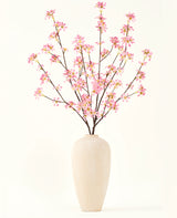 En hög beige keramikvas innehåller flera grenar från Silk konstgjorda blombuketten Rosa blommor, med fina rosa blommor och gröna blad, mot en vanlig vit bakgrund.