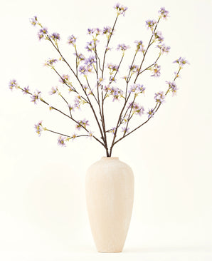 En hög, krämfärgad keramikvas innehåller ett antal tunna grenar med texten Silk konstgjord blombukett Konstgjord blomgren lila, mot en slät, ljus bakgrund.