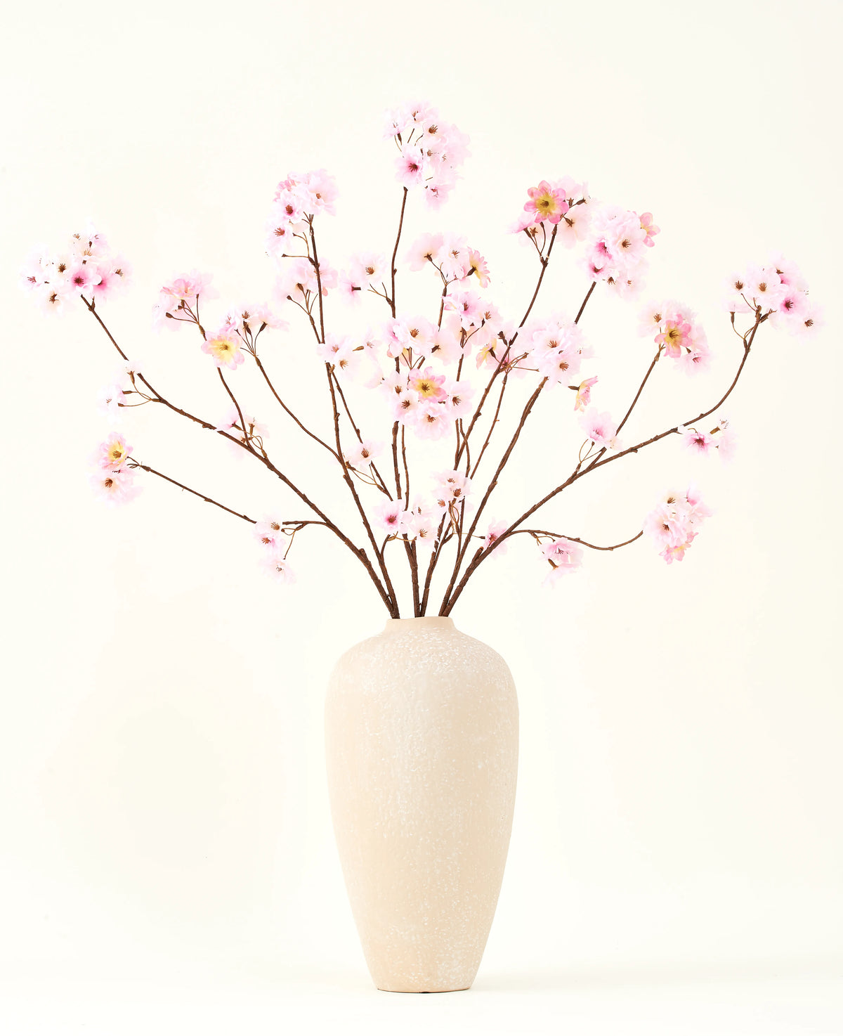 Siden konstgjorda blombuketten Körsbärsblomrosa 114 cm har långa tunna grenar med klasar av ljusrosa blommor i en beige keramikvas, som bildar en slående kontrast mot den enkla vita bakgrunden.