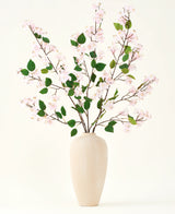 En beige keramikvas innehåller flera långa grenar med Silk artificiell blombukett av Peach Blossom rosa och gröna blad, mot en vanlig vit bakgrund - perfekt för en konstgjord blommig inredning.