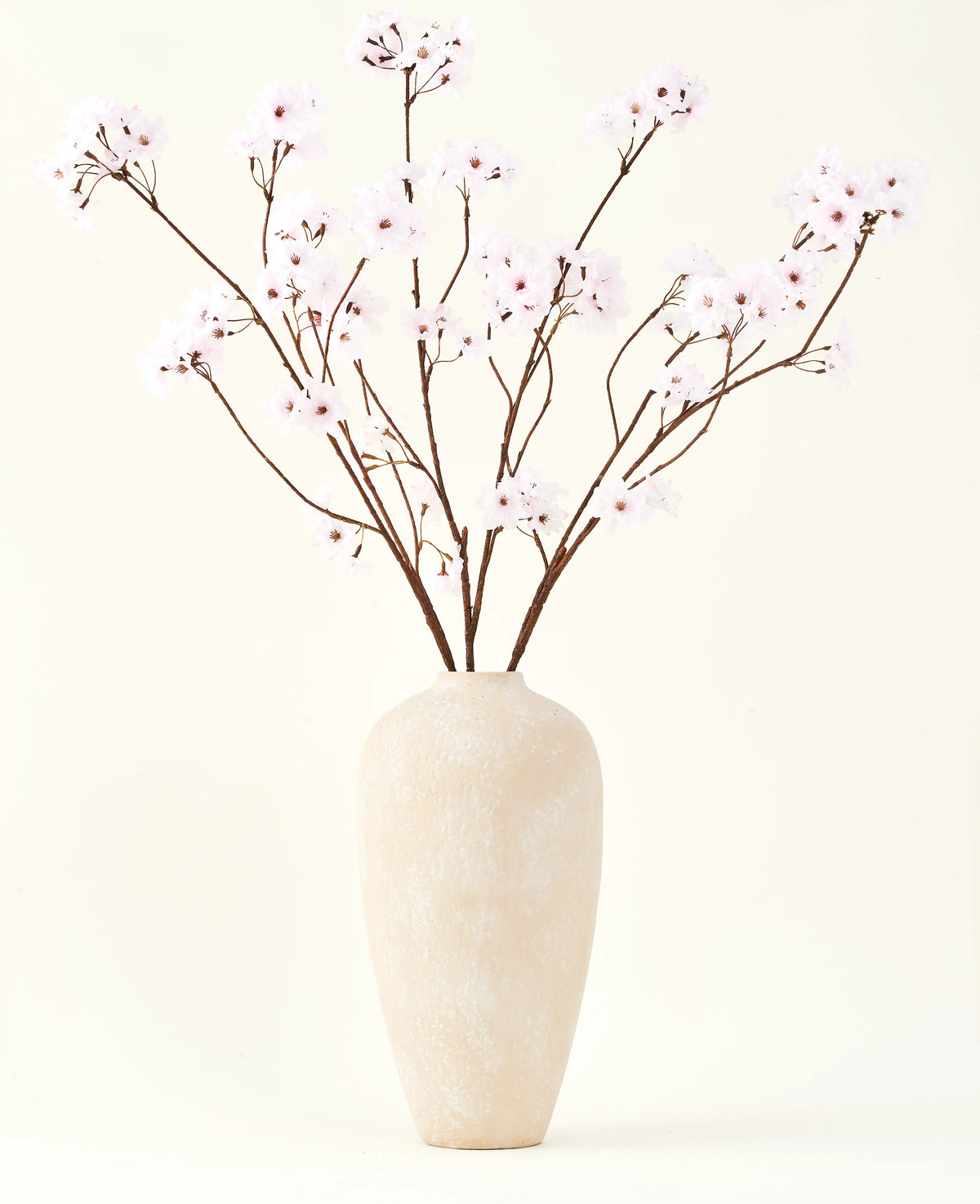 Sidens konstgjorda blombukett Körsbärsblommor ljusrosa 114 cm står i en hög beige keramikvas med långa, tunna grenar och delikata ljusrosa blommor, vilket skapar en fridfull konstgjord blommig accent mot en enkel bakgrund.
