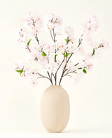 Sidens konstgjorda blombukett Körsbärsblomma rouge 119 cm visar delikata, ljusrosa körsbärsblommor med gröna blad i en beige vas, perfekt som en konstgjord blomdekoration mot en ljus bakgrund.