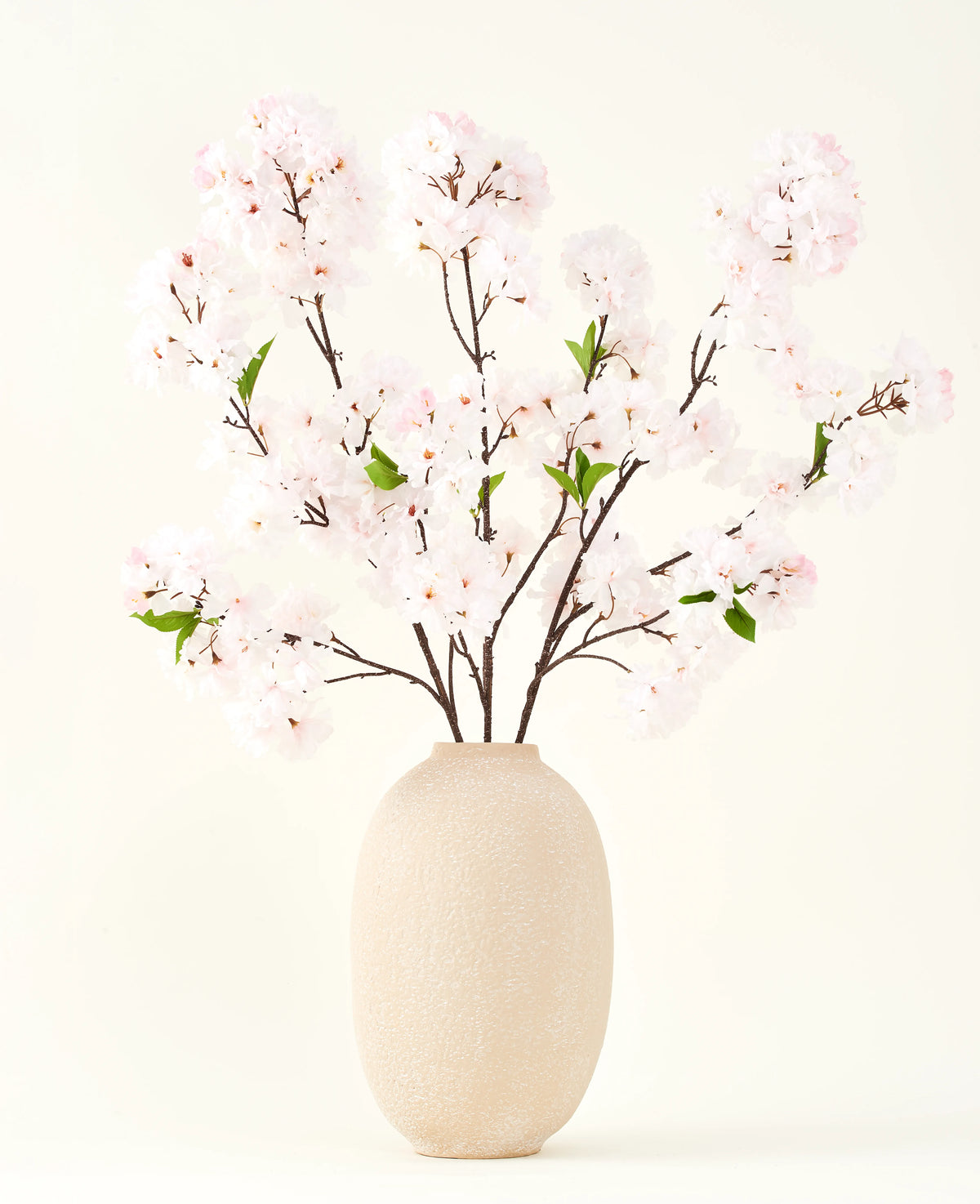 Sidens konstgjorda blombukett Körsbärsblomma rouge 119 cm visar delikata, ljusrosa körsbärsblommor med gröna blad i en beige vas, perfekt som en konstgjord blomdekoration mot en ljus bakgrund.