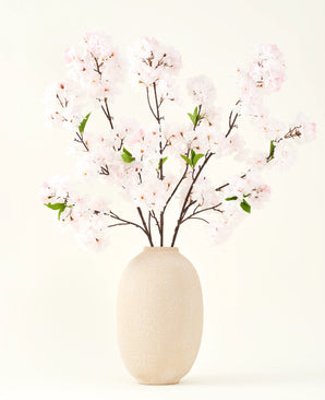 Sidens konstgjorda blombukett Körsbärsblomma rouge 119 cm visar delikata, ljusrosa körsbärsblommor med gröna blad i en beige vas, perfekt som en konstgjord blomdekoration mot en ljus bakgrund.