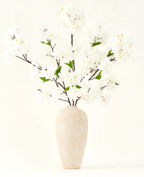 Silk konstgjorda blombuketten Cherry blossom cream 119 cm har eleganta bruna grenar med mjuka vita falska blommor och gröna blad, utställd i en enkel beige vas mot en enkel ljus bakgrund.
